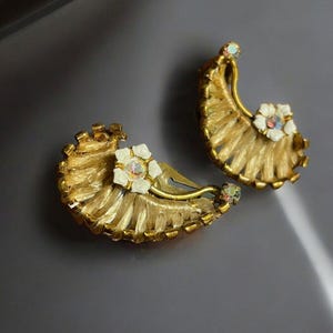 May include: A pair of gold tone clip-on earrings with a woven design and white flower accents. The flowers have iridescent rhinestones in the center.