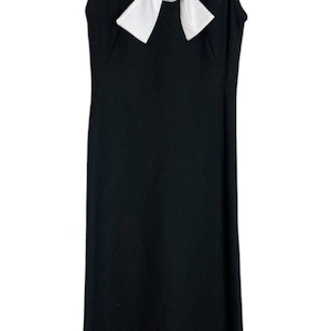 Puede incluir: Vestido negro sin mangas con cuello y lazo blancos. El cuello tiene un patrón texturizado con detalles florales negros. El vestido está colgado en una percha negra.