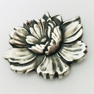 May include: A silver flower brooch with a detailed, textured design. The flower has multiple petals and a center with a raised, textured center.