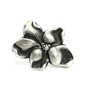 Puede incluir: Un broche de plata con forma de flor con un dise&ntilde;o detallado y texturizado. La flor tiene cinco p&eacute;talos y un centro con un peque&ntilde;o detalle en relieve.