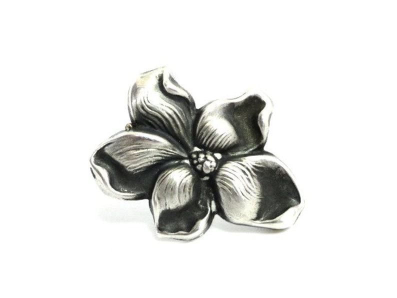 Puede incluir: Un broche de plata con forma de flor con un dise&ntilde;o detallado y texturizado. La flor tiene cinco p&eacute;talos y un centro con un peque&ntilde;o detalle en relieve.