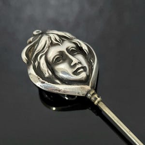 May include: A silver-toned hat pin featuring a detailed face design. The face has flowing hair and a serene expression. The pin has a long, slender stem and a decorative head.