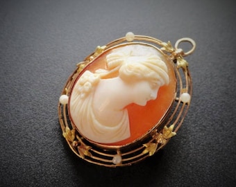 Puede incluir: Un colgante de camafeo de oro con un retrato tallado de una mujer con un vestido blanco. El camafeo está engastado en un marco de oro con un delicado diseño floral y detalles de perlas.