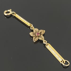 Op de afbeelding: Een gouden armbandonderdeel met een centrale vijfbladige bloem met een kleine rode steen in het midden. De bloem wordt geflankeerd door twee langwerpige, getextureerde gouden staven, met een veerringsluiting aan het ene uiteinde en een ring.