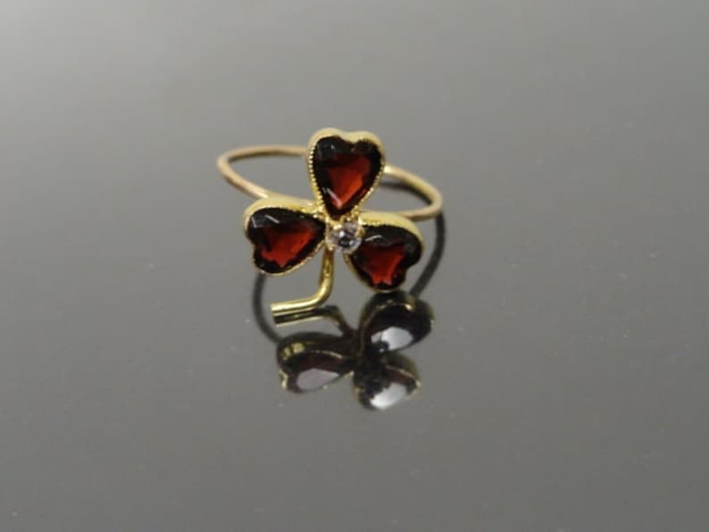May include: A gold ring with a three-leaf clover design. Each leaf is set with a red gemstone, and a smaller white gemstone is in the center.