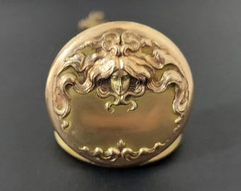 May include: A round gold-toned metal compact case featuring an embossed relief of a face with long, flowing hair that extends across the upper portion of the lid. The surface has a polished finish with intricate decorative scrollwork at the base.