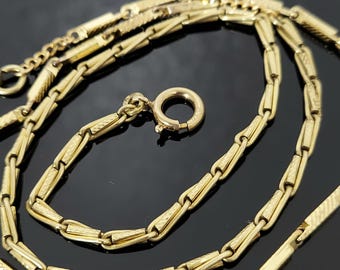 Puede incluir: Un collar dorado con un diseño de eslabones único. El collar presenta una serie de eslabones rectangulares y estampados, conectados por segmentos más pequeños y texturizados. Se ve un cierre circular, y la cadena se muestra sobre un fondo oscuro.