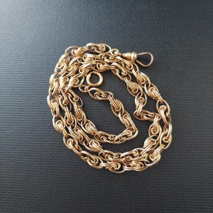 May include: A gold-coloured chain necklace with a clasp and a circular ring. The chain is made of multiple interlocking links, creating a textured appearance. The necklace is displayed on a dark background.