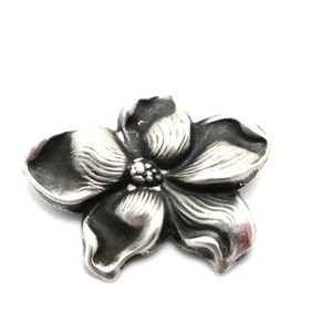 Puede incluir: Un amuleto de flor de plata con un dise&ntilde;o detallado. La flor tiene cinco p&eacute;talos y un centro con un peque&ntilde;o c&iacute;rculo en relieve.