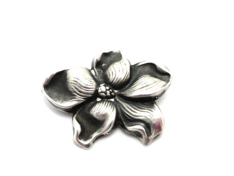 Puede incluir: Un amuleto de flor de plata con un dise&ntilde;o detallado. La flor tiene cinco p&eacute;talos y un centro con un peque&ntilde;o c&iacute;rculo en relieve.