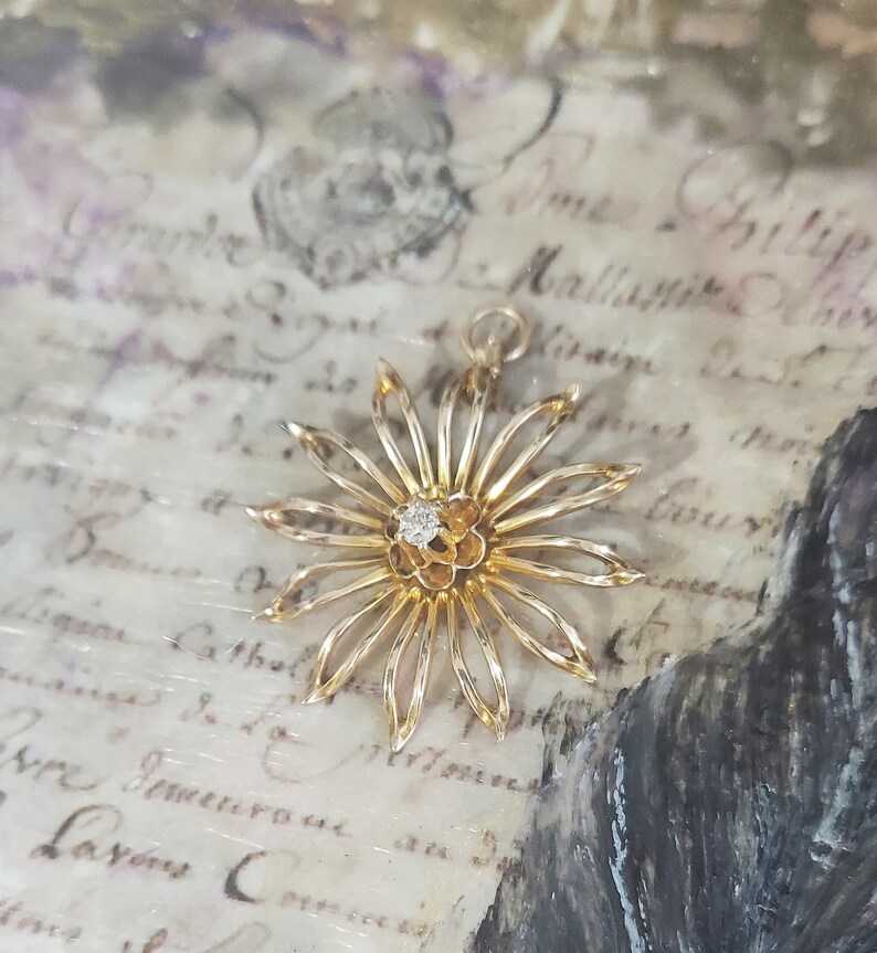 May include: A gold-toned flower pendant with a single diamond in the centre. The pendant is on a white background with cursive writing.