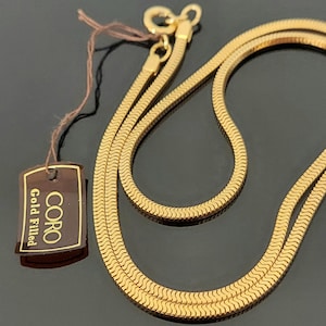 Puede incluir: Un collar de cadena de espiga relleno de oro con un cierre de resorte. Una etiqueta rectangular marrón está unida al collar por un hilo fino, con el texto CORO Gold Filled en letras doradas sobre un fondo marrón oscuro.