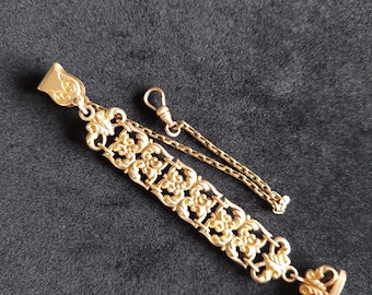 Op de afbeelding: Een gouden zakhorlogeketting met een decoratief, versierd ontwerp. De ketting heeft aan het ene uiteinde een sluiting en aan het andere uiteinde een hanger.