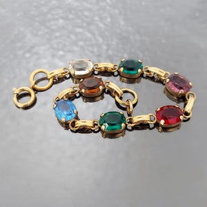 Pode incluir: Pulseira dourada com uma série de pedras preciosas ovais coloridas. As pedras são engastadas em biséis individuais e unidas por pequenos conectores dourados. A pulseira inclui um fecho para uso seguro.