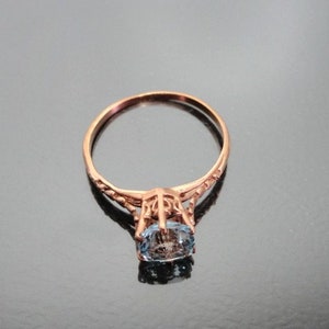 K&ouml;nnte beinhalten: Ein zarter Ring aus Ros&eacute;gold mit einem ovalen blauen Edelstein, der in einem filigranen Design gefasst ist.