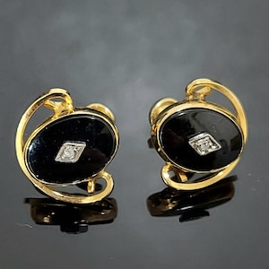 Puede incluir: Un par de pendientes dorados con piedras de ónix negro ovaladas y un pequeño diamante en el centro. Los pendientes tienen un diseño curvo único y se muestran sobre una superficie reflectante.