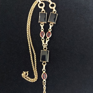 Puede incluir: Un llavero de cadena de reloj de oro con detalles en negro y rojo. El llavero tiene un cierre en forma de T y una cadena con piedras rectangulares negras y ovaladas rojas.