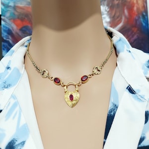 May include: A gold necklace with a heart-shaped pendant and red gemstones. The pendant has a detailed design and a small red gemstone in the center.