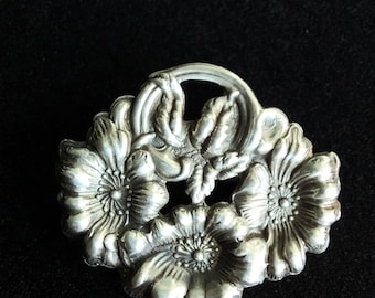 May include: A silver brooch with a floral design. The brooch features three stylized flowers with detailed petals and leaves.