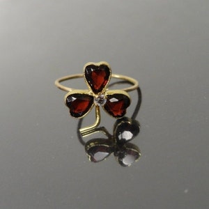 May include: A gold ring with a three-leaf clover design. Each leaf is set with a dark red gemstone. A small clear gemstone is in the center of the clover.