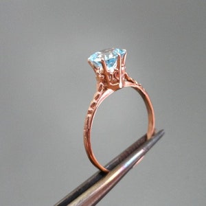 K&ouml;nnte beinhalten: Ein Ring aus Ros&eacute;gold mit einem hellblauen Edelstein, der in einer zarten, verzierten Fassung sitzt.