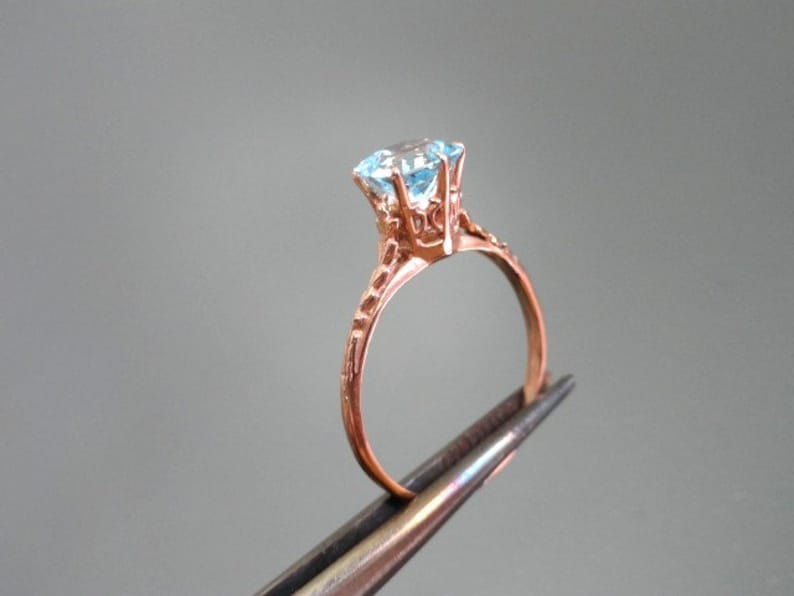 K&ouml;nnte beinhalten: Ein Ring aus Ros&eacute;gold mit einem hellblauen Edelstein, der in einer zarten, verzierten Fassung sitzt.