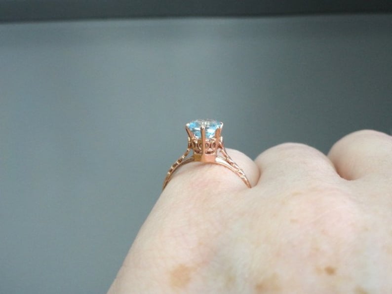 K&ouml;nnte beinhalten: Ein Verlobungsring aus Ros&eacute;gold mit einem gro&szlig;en, klaren blauen Edelstein, der in einer filigranen, verzierten Fassung sitzt.