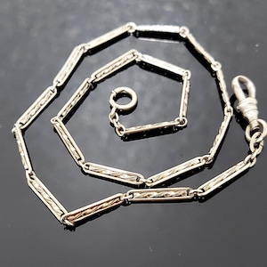 May include: A silver-toned watch chain with a decorative pattern. The chain features rectangular links with an etched design, connected by smaller links. It has a clasp on one end and a ring on the other, suitable for attaching to a pocket watch.