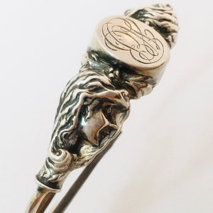 May include: Silver serving tongs with an intricate design. The tongs feature a sculpted female face and a circular top with an engraved monogram. The handle is curved and the overall design is ornate.