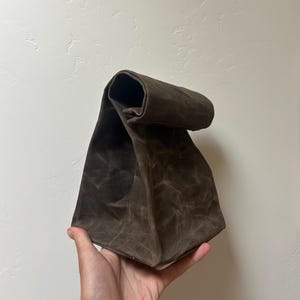 May include: A dark brown, waxed canvas lunch bag with a roll-top closure. The bag features a structured, rectangular base and a textured, weathered appearance characteristic of waxed fabric, with visible creases and a matte finish throughout.