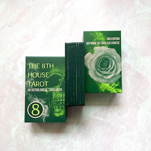 May include: A green tarot deck box with a skull and the text "The 8th House Tarot, An Astrological Tarot Deck" and the number "8" in a circle. The box is sitting on a white surface. The back of the box is visible, showing the text "3rd Edition, Artwork by Sara Calvarese" and a white rose with green smoke.