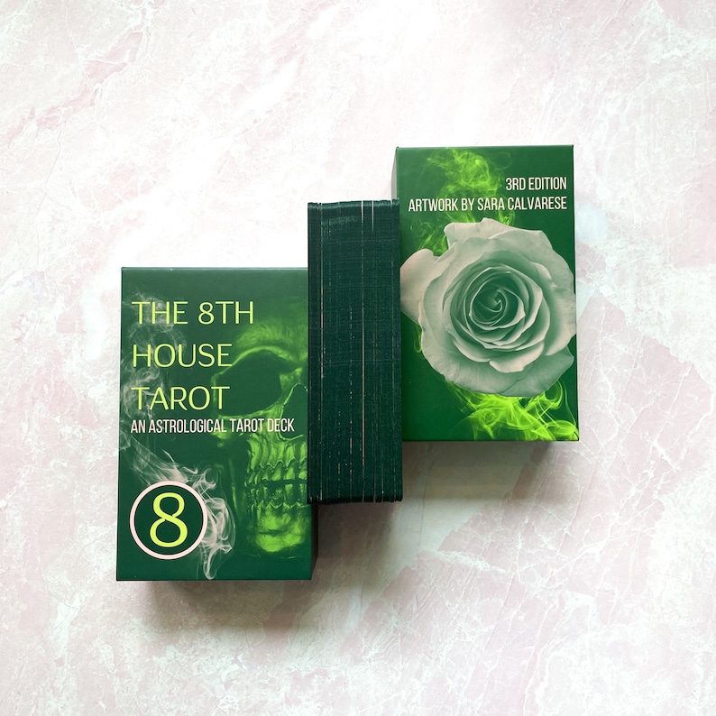 May include: A green tarot deck box with a skull and the text "The 8th House Tarot, An Astrological Tarot Deck" and the number "8" in a circle. The box is sitting on a white surface. The back of the box is visible, showing the text "3rd Edition, Artwork by Sara Calvarese" and a white rose with green smoke.