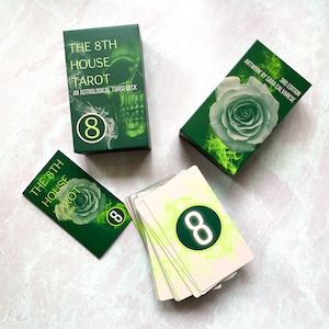 May include: The 8th House Tarot deck box features a green skull with a yellow number 8. The box is on a light pink surface. The deck is an astrological tarot deck. The 3rd edition of the deck is by Sara Calvarese.