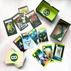 May include: A set of tarot cards with green and yellow accents. The cards feature illustrations of various symbols and imagery. The cards are arranged in a circle around a box with the number 8 on it.