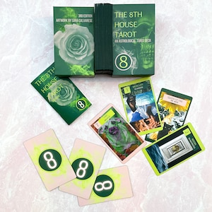 May include: A green tarot deck with the title "The 8th House Tarot: An Astrological Tarot Deck" and a skull graphic. The deck is laid out on a pink surface with several cards from the deck, including the Queen of Fire, the Moon, and the Magician.