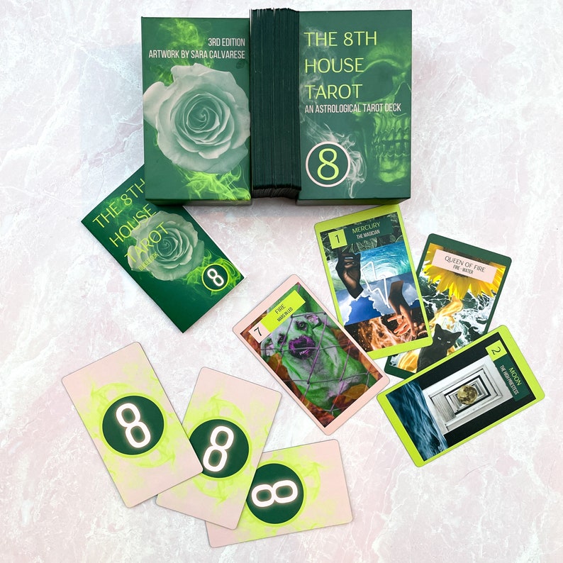 May include: A green tarot deck with the title "The 8th House Tarot: An Astrological Tarot Deck" and a skull graphic. The deck is laid out on a pink surface with several cards from the deck, including the Queen of Fire, the Moon, and the Magician.