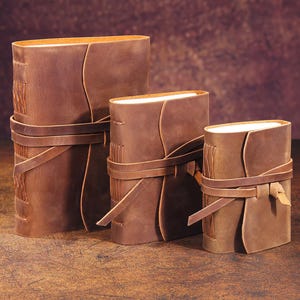 May include: Three brown leather-bound journals of varying sizes, each with a leather strap closure. The journals are arranged in descending order of size, with the largest journal on the left and the smallest journal on the right.