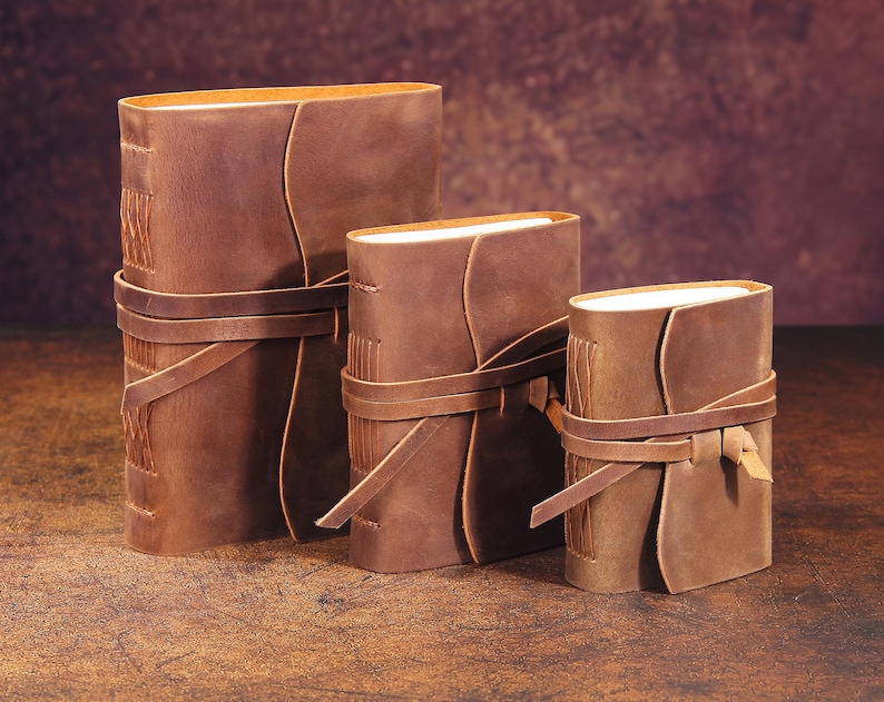 May include: Three brown leather-bound journals of varying sizes, each with a leather strap closure. The journals are arranged in descending order of size, with the largest journal on the left and the smallest journal on the right.