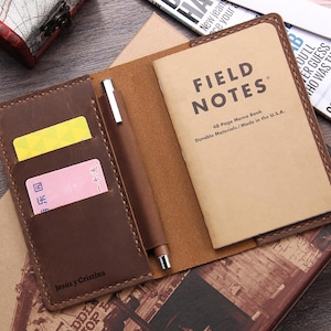 May include: A brown leather notebook cover with a pen holder and two card slots. The cover is personalized with the text "Jesús y Cristina". The notebook inside is a Field Notes brand memo book with the text "Field Notes 48-Page Memo Book Durable Materials/Made in the U.S.A."