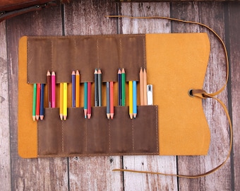 May include: A brown leather pencil roll with a yellow flap and a strap. The roll is open and contains a variety of colored pencils.