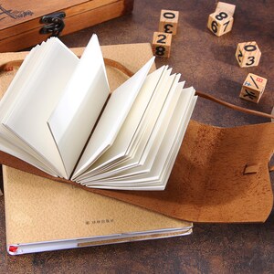 May include: A brown leather-bound journal with a blank page open. The journal is lying on a table with a wooden box and wooden number blocks nearby.