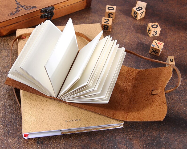 May include: A brown leather-bound journal with a blank page open. The journal is lying on a table with a wooden box and wooden number blocks nearby.