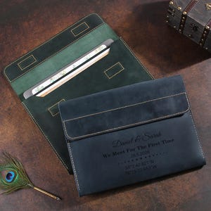 May include: A dark blue leather document folder with a flap closure, engraved with the text Daniel & Sarah, We Meet For The First Time, 28.5.2026, and coordinates. A second green leather folder is partially visible underneath the blue one.