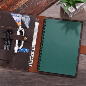 Peut inclure: Un journal en cuir marron avec un carnet vert et plusieurs poches. Le journal contient des stylos, un câble de chargement et des cartes. Le texte "I miss you more -MK" est visible. Fermeture à bouton-pression.