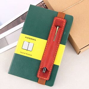May include: A green notebook with a yellow and white label that says "PAPERERA Squared Notebook". A red leather pen holder with a gold pen is attached to the notebook.