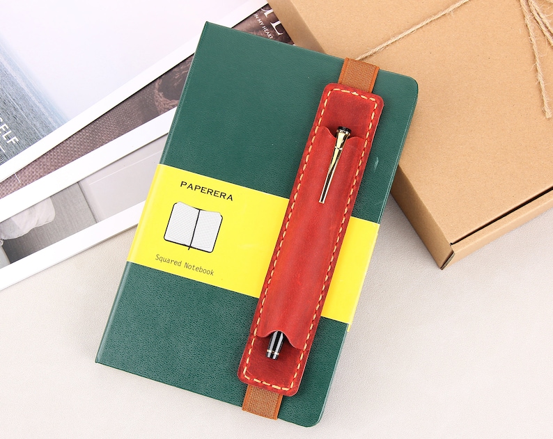 May include: A green notebook with a yellow and white label that says "PAPERERA Squared Notebook". A red leather pen holder with a gold pen is attached to the notebook.