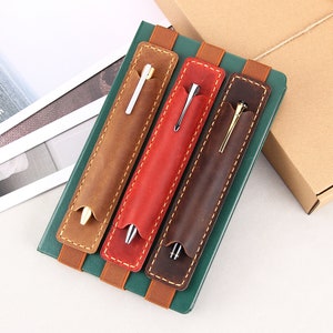 May include: Three leather pen holders with elastic bands, each holding a different coloured pen. The holders are brown, red, and dark brown.