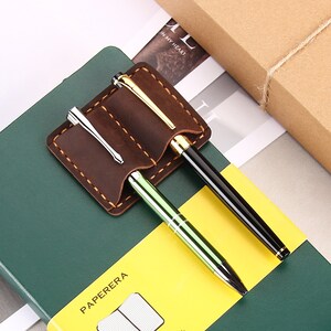 May include: A brown leather pen holder with two slots for pens. The holder is attached to a green notebook with the word "PAPERERA" printed on the cover.