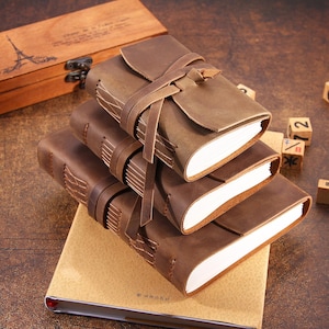 May include: Three brown leather journals with a rustic look, stacked on top of each other. The top journal has a leather strap closure and is wrapped in a brown leather cover. The journals are on a beige surface.
