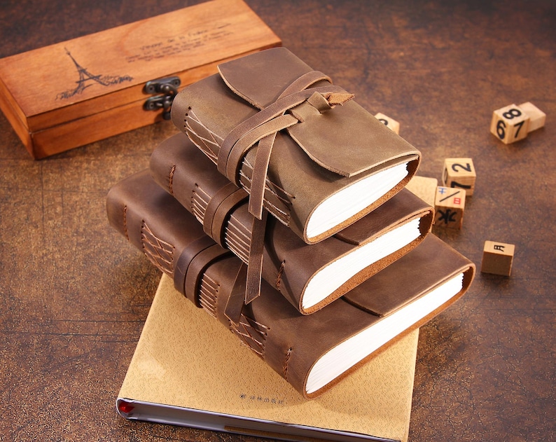 May include: Three brown leather journals with a rustic look, stacked on top of each other. The top journal has a leather strap closure and is wrapped in a brown leather cover. The journals are on a beige surface.